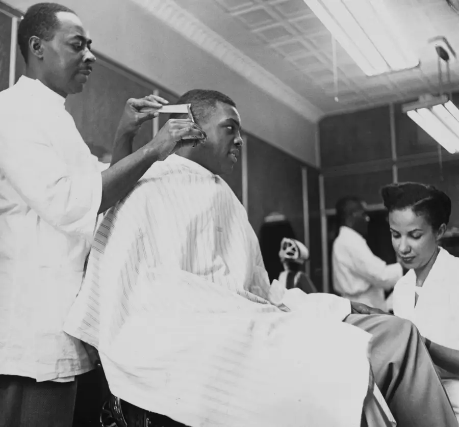 A barber trims a man’s hair while a woman sits nearby, both wearing white uniforms, in a barbershop with striped capes and vintage lighting overhead.