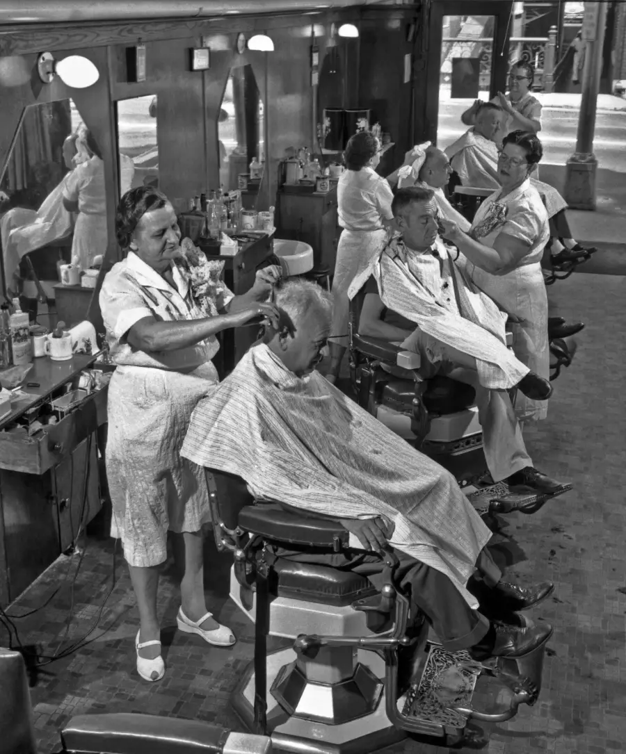 Black-and-white photo of three men getting haircuts from three women in a vintage barbershop, with mirrors, barber chairs, and supplies visible. Sunlight streams through large windows in the background.
