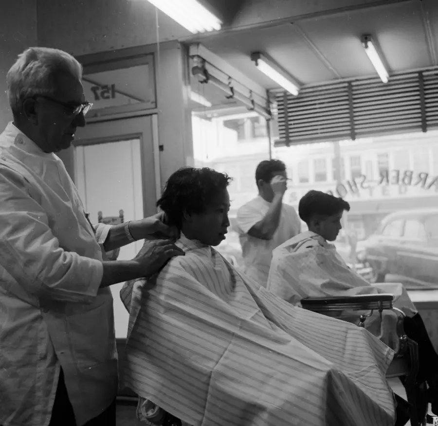 A barber stands beside a boy in a striped cape, preparing to cut his hair. Another barber works with a customer in the background. The scene is inside a barbershop with large front windows.