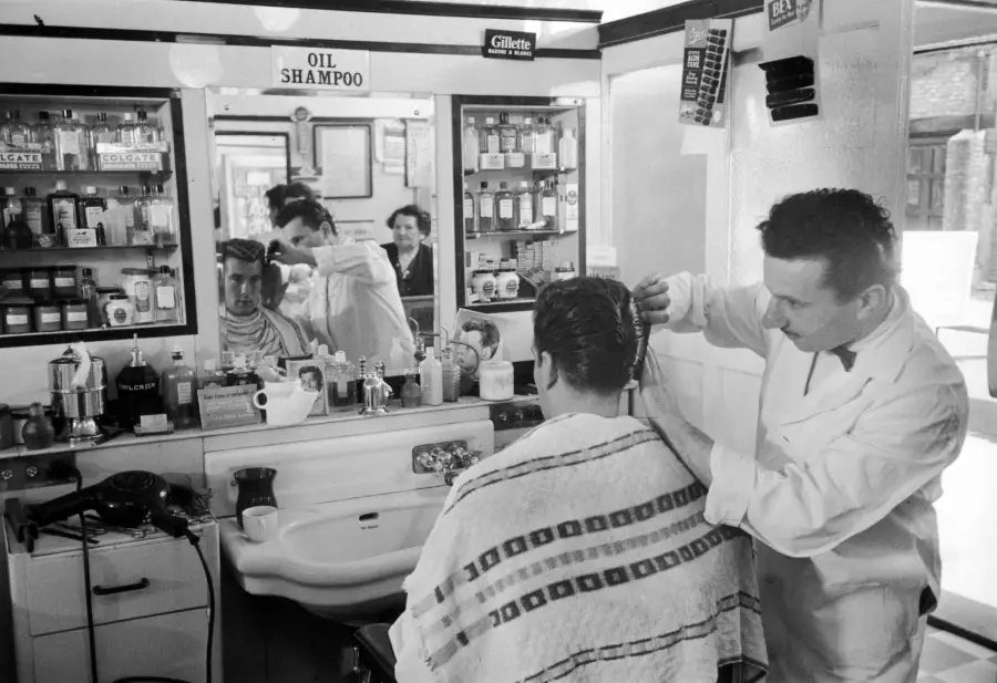 A barber wearing a white coat cuts a customer’s hair in a vintage barbershop, with shelves of products and a mirror reflecting both men.