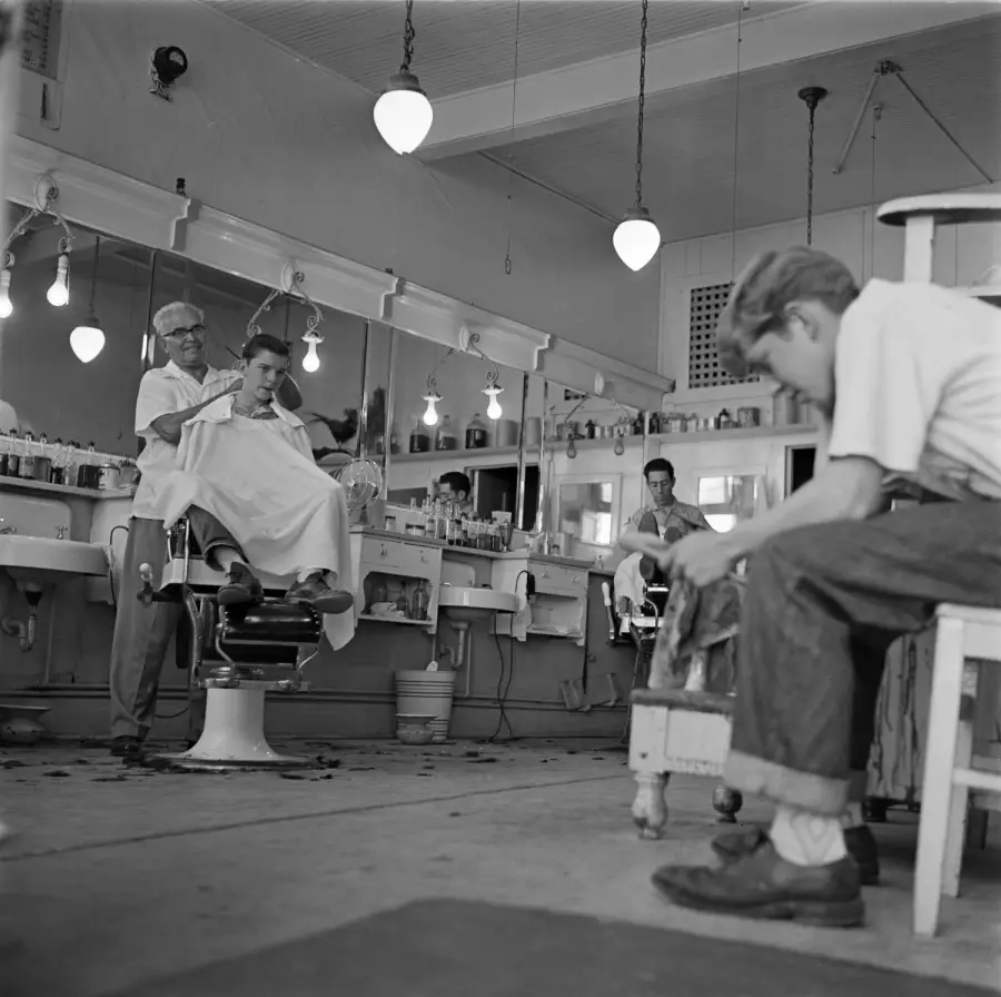 A barber cuts a customer’s hair in a vintage barbershop. Mirrors, lightbulbs, and chairs line the wall. Another barber works in the background, while a boy sits in the foreground, looking down.