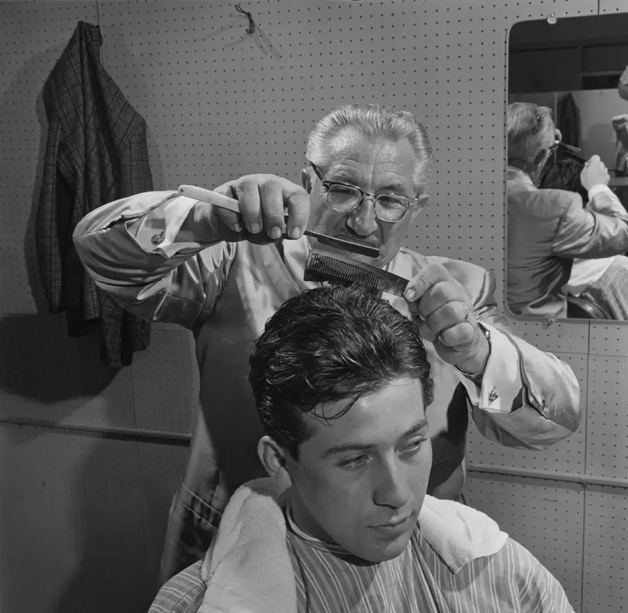 A barber combs and cuts a young man’s hair in a barbershop. Both are focused on the haircut. A jacket hangs on the wall, and their reflection is visible in a mirror behind them.