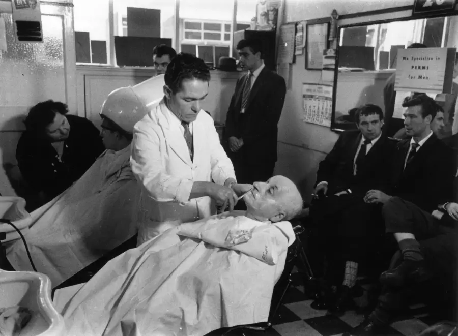 A barber shaves a man’s face while others wait and watch in a busy barbershop. Some customers are seated in barber chairs; others wait against the wall. The scene is indoors and appears to be from the mid-20th century.