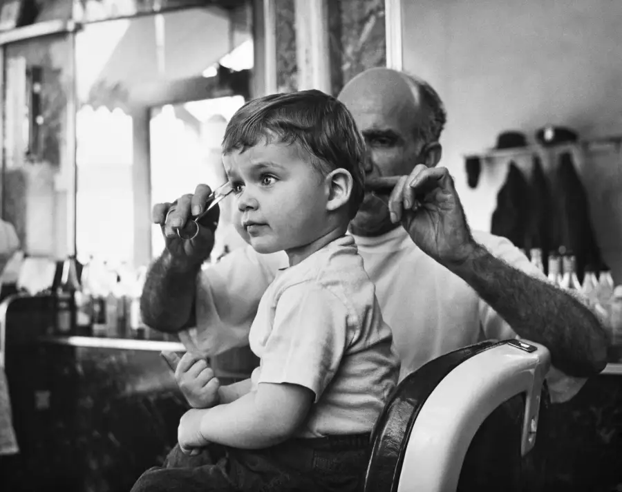 A young boy sits in a barber’s chair, looking ahead, while a barber with scissors prepares to cut his hair. The setting appears to be an old-fashioned barbershop.