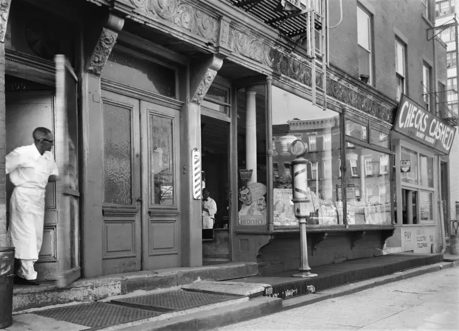 Black-and-white photo of a man exiting a shop with ornate trim. Next door is a store with a “Checks Cashed” sign and goods in the window. Another man is visible inside, seated near a barber pole.