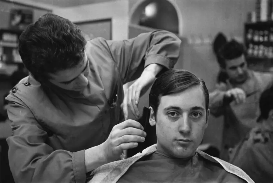 A young man gets a haircut at a barbershop. The barber carefully trims around his ear while another person is seen getting a haircut in the background.