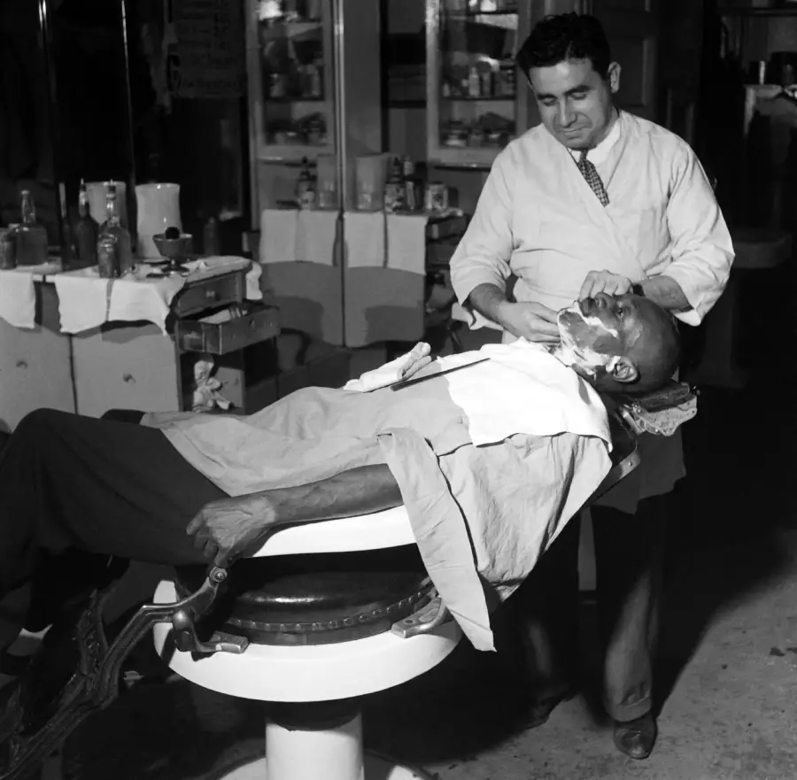 A barber shaves a man who is reclining in a vintage barber chair. The barber is focused and the client is relaxed, with towels around his neck. Bottles and supplies are visible in the background.