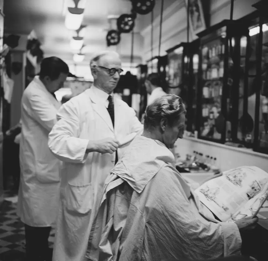 A black-and-white photo of a barber cutting a man’s hair while the customer reads a newspaper. Another barber is working in the background. Both barbers wear white coats. The setting appears to be a vintage barbershop.