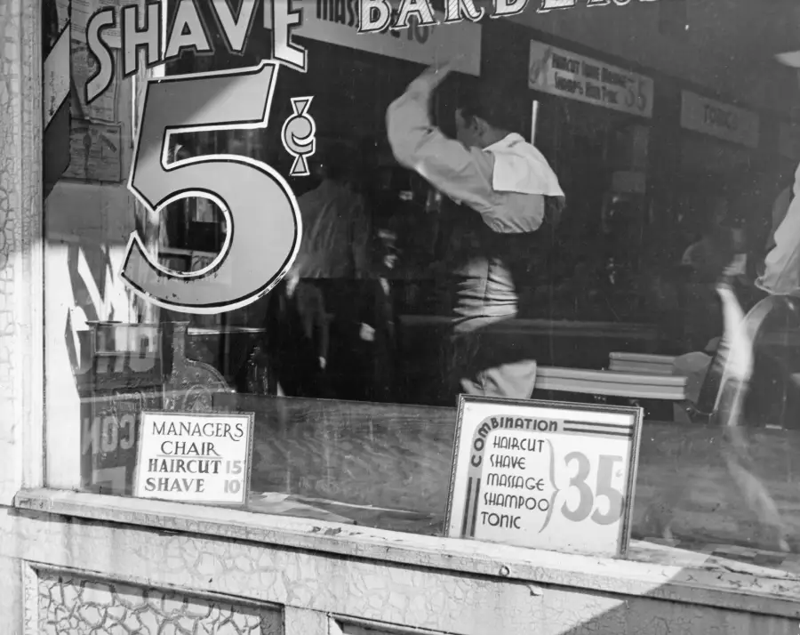 Black-and-white photo of a barber shop window advertising shaves for 5 cents. Price lists show haircuts for 15 cents and combination services for 35 cents. A barber is seen working inside.