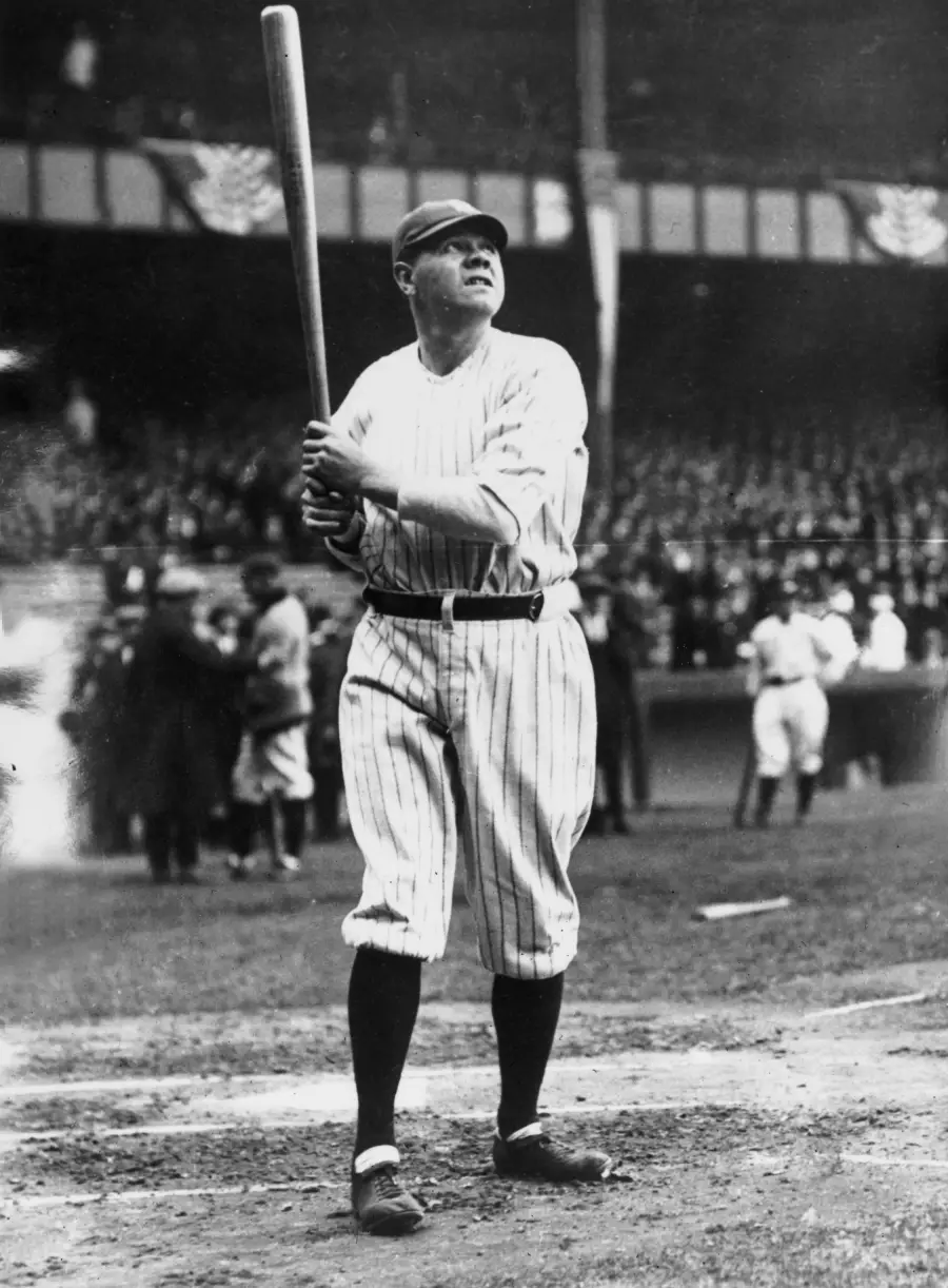 Baseball player in pinstripe uniform holding a bat, standing at home plate on a field, with spectators and other players visible in the background. The image is black and white.