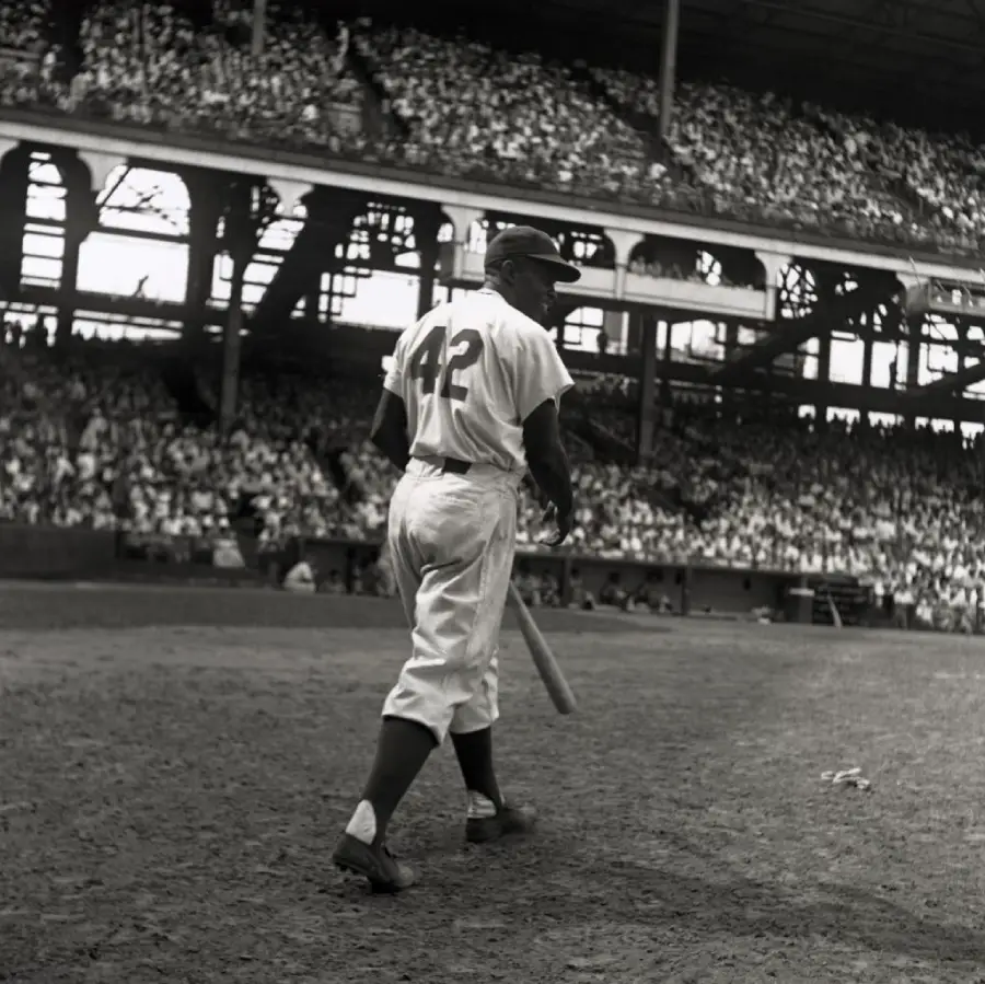 A baseball player wearing a jersey with the number 42 walks onto the field holding a bat, with a packed stadium and cheering crowd in the background.