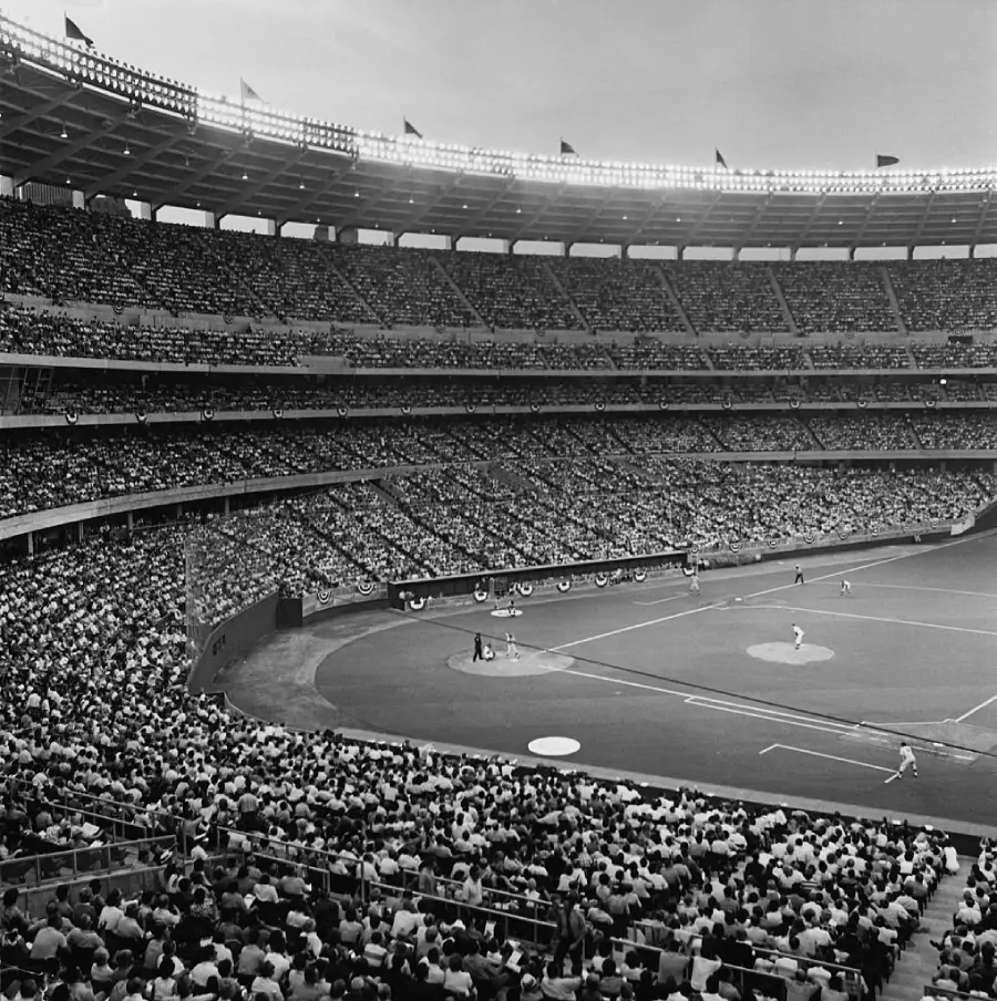 A packed baseball stadium filled with fans watching a game in progress, with players on the field and bright stadium lights illuminating the scene.