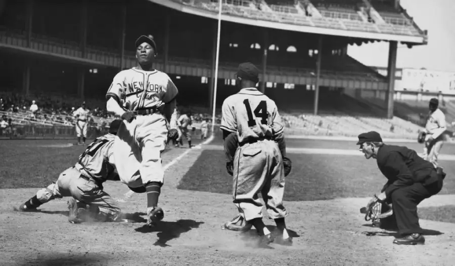 A baseball player in a “New York” uniform scores at home plate as the catcher and another player look on, with an umpire crouched nearby in a large stadium.