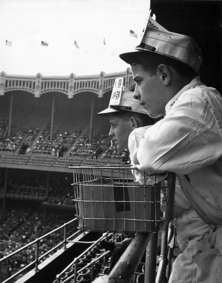 Two young men in vendor uniforms and hats lean on a railing, overlooking a crowded baseball stadium. One has a metal basket for goods. The scene is black and white and appears to be from the mid-20th century.