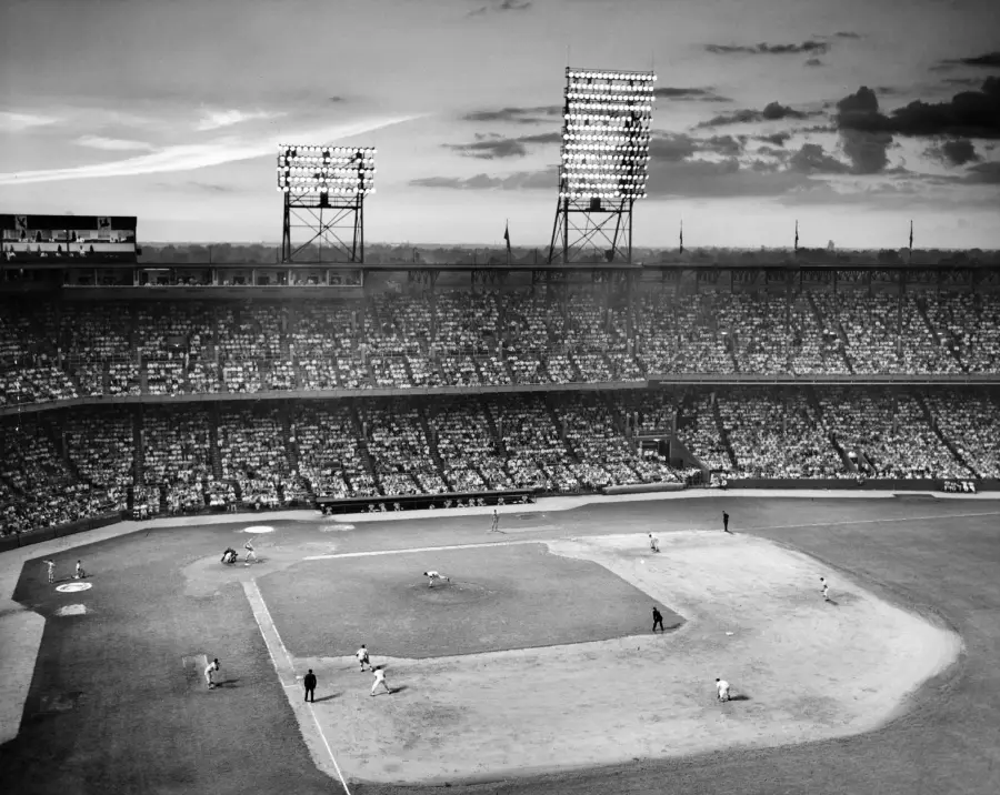 A black and white photo of a crowded baseball stadium during a night game, with players on the field and bright stadium lights illuminating the scene.