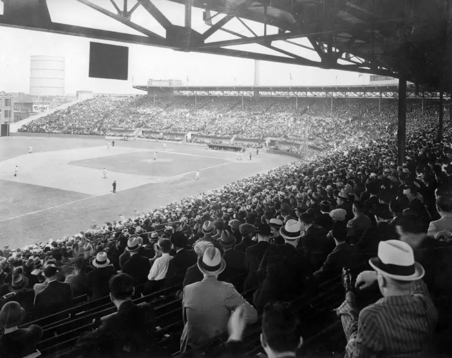 Black-and-white photo of a packed baseball stadium in the early 20th century, with spectators in hats watching a game on the field from covered grandstand seats.