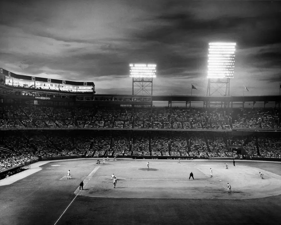 A black-and-white photo of a crowded baseball stadium at night, brightly lit by floodlights, with players on the field and fans filling the stands.