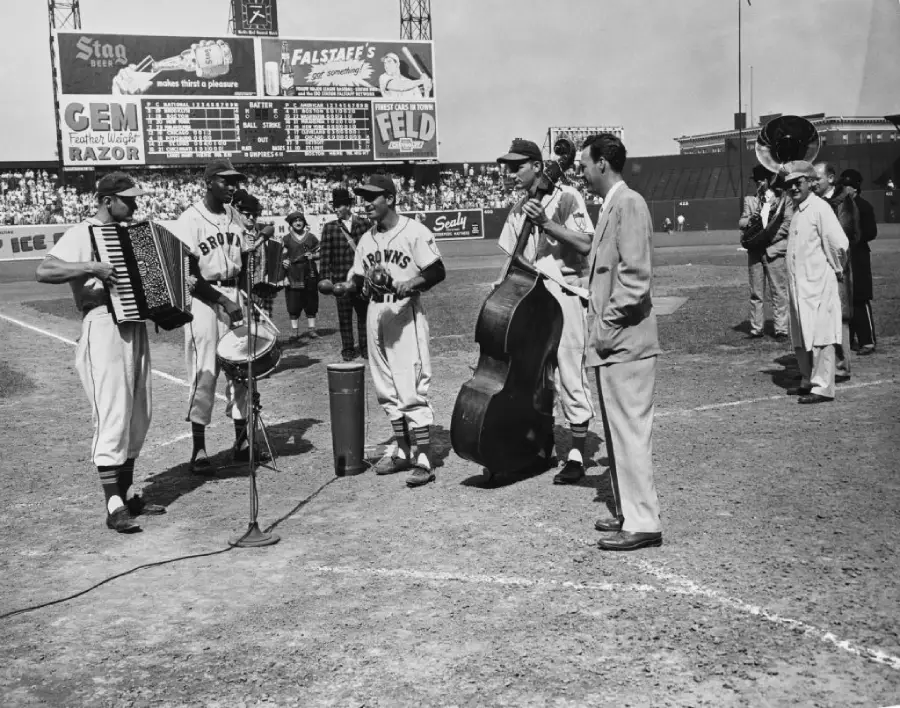 Black and white photo of baseball players in uniforms playing instruments on a baseball field. A scoreboard, crowd, and several people, including a man in a suit, are visible in the background.