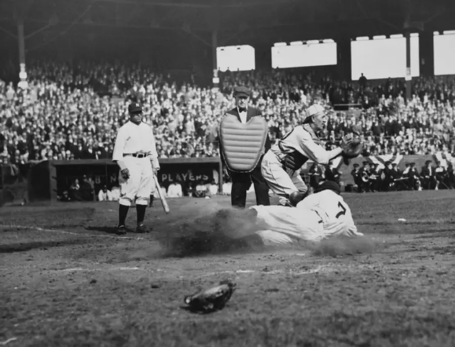 A baseball player slides into home plate as the catcher tries to tag him. An umpire watches closely, while another player stands nearby. The crowd fills the stadium seats in the background.