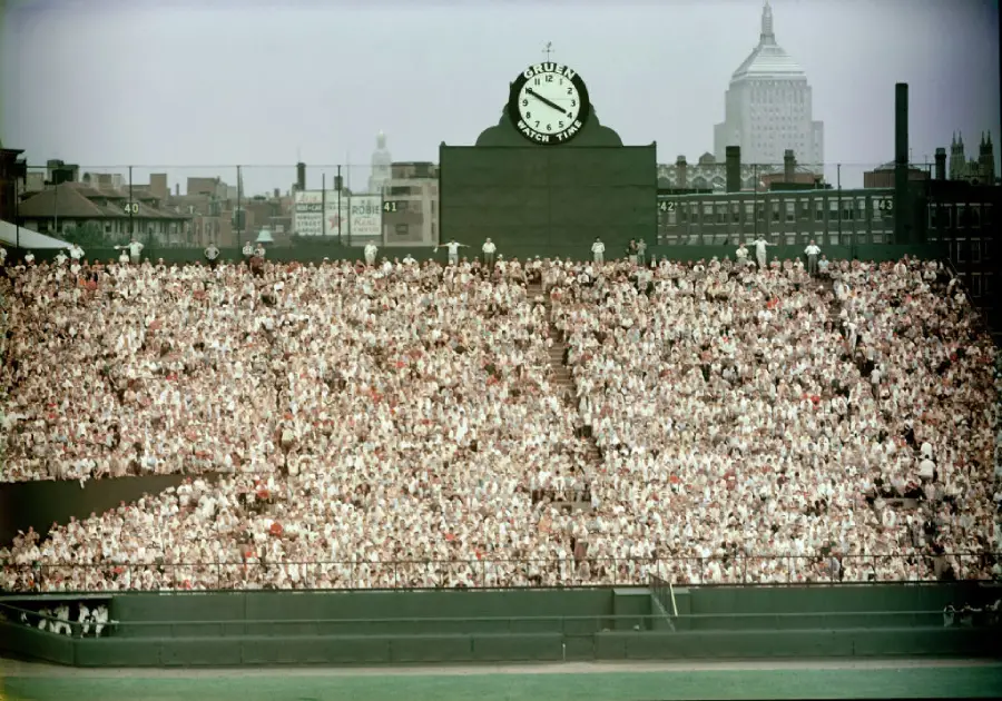 A large crowd fills Fenway Park’s bleachers on a sunny day in front of the iconic center field clock, with Boston city buildings visible in the background.