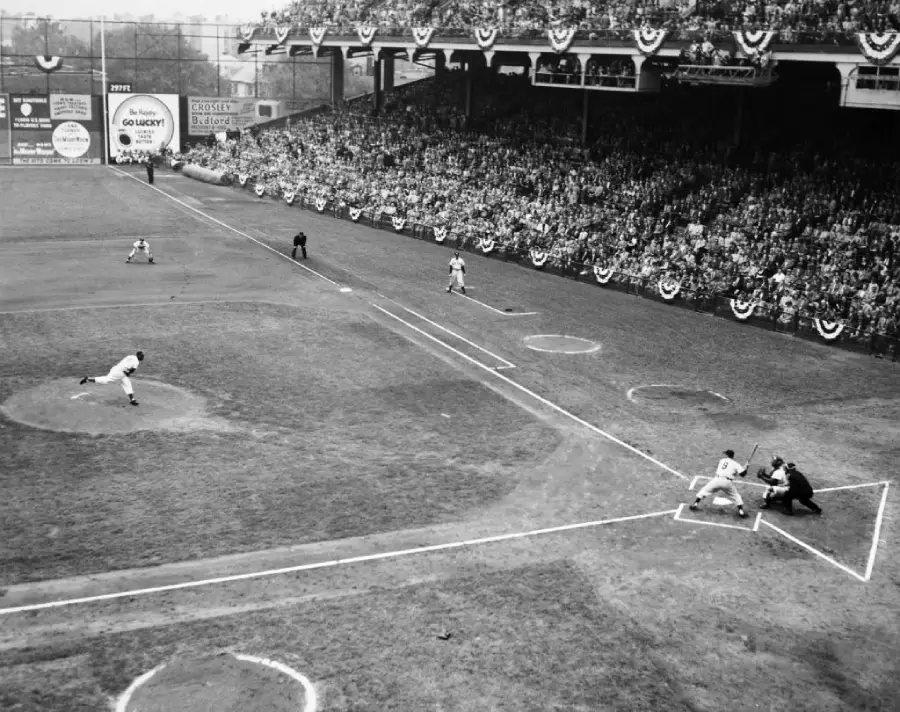Black-and-white photo of a baseball game in a crowded stadium, showing the pitcher throwing to the batter, with fans filling the stands decorated with banners.