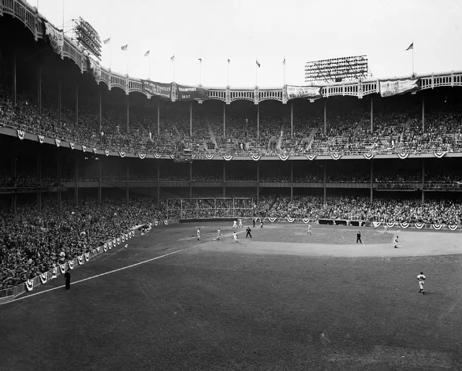Black and white photo of a crowded baseball stadium during a game, with spectators filling the stands and flags flying above the grandstand. The field is decorated with bunting.