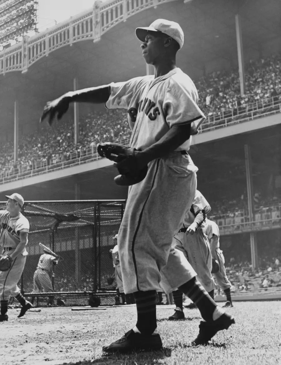 Black-and-white photo of a baseball player wearing a Giants uniform, throwing a ball on a field, with teammates and a crowded stadium visible in the background.