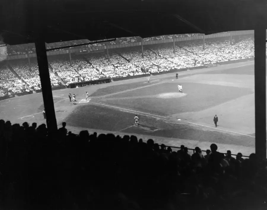 Black and white photo of a crowded baseball stadium, with players on the field and spectators filling the stands. The image is taken from a shaded area behind home plate.