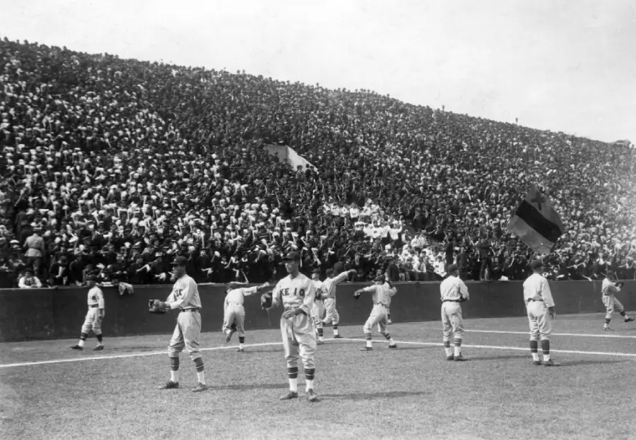 Baseball players warm up on the field while a large crowd fills the stadium seats behind them, watching the game intently.