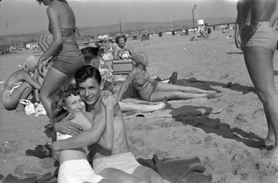 A man and a girl smile and hug while sitting on a sandy beach, surrounded by people sunbathing and relaxing. Swimmers and sunbathers, both adults and children, are scattered along the beach on a sunny day.