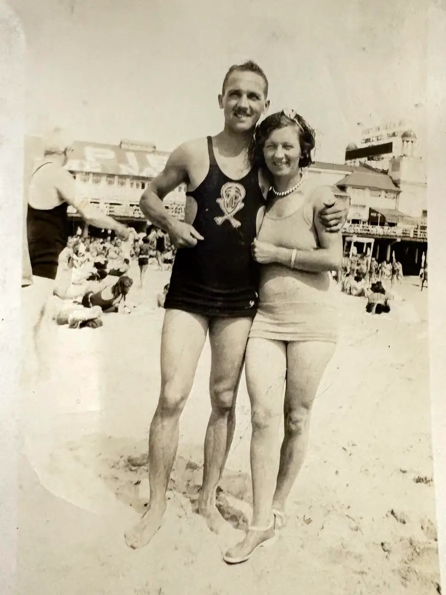 A smiling man and woman in vintage swimsuits stand arm-in-arm on a crowded beach, with buildings and other beachgoers visible in the background.