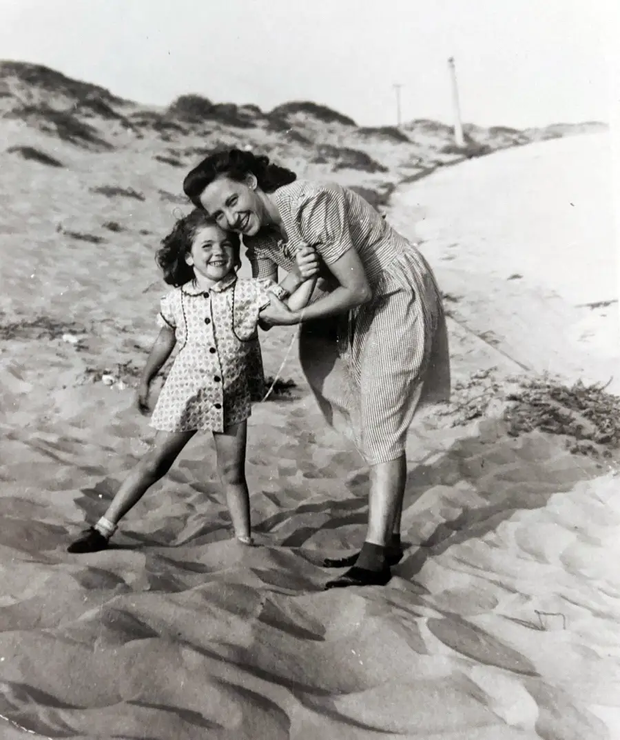A woman and a young girl smile while standing together on a sandy beach, surrounded by dunes. Both wear patterned dresses and dark shoes. The girl leans playfully as they embrace. The photo is black and white.