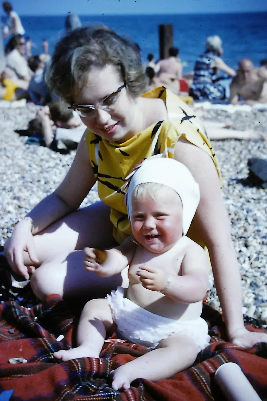 A woman in a yellow dress sits on a beach blanket with a smiling baby wearing a white bonnet and diaper, with other people and the sea in the background.