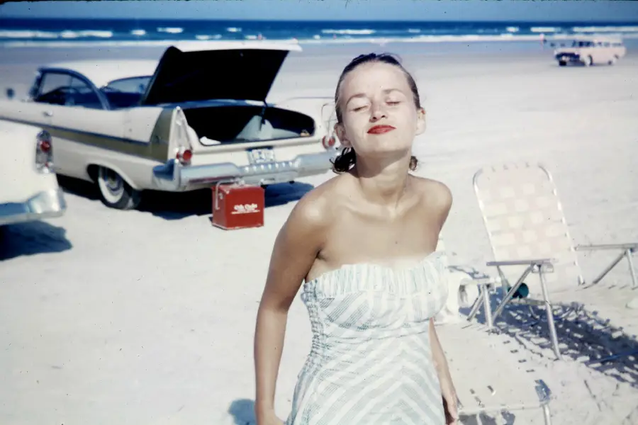 A woman in a strapless dress smiles with closed eyes on a sandy beach. Behind her, a classic car with an open trunk and a cooler is parked. The ocean and beach chairs are visible in the background.