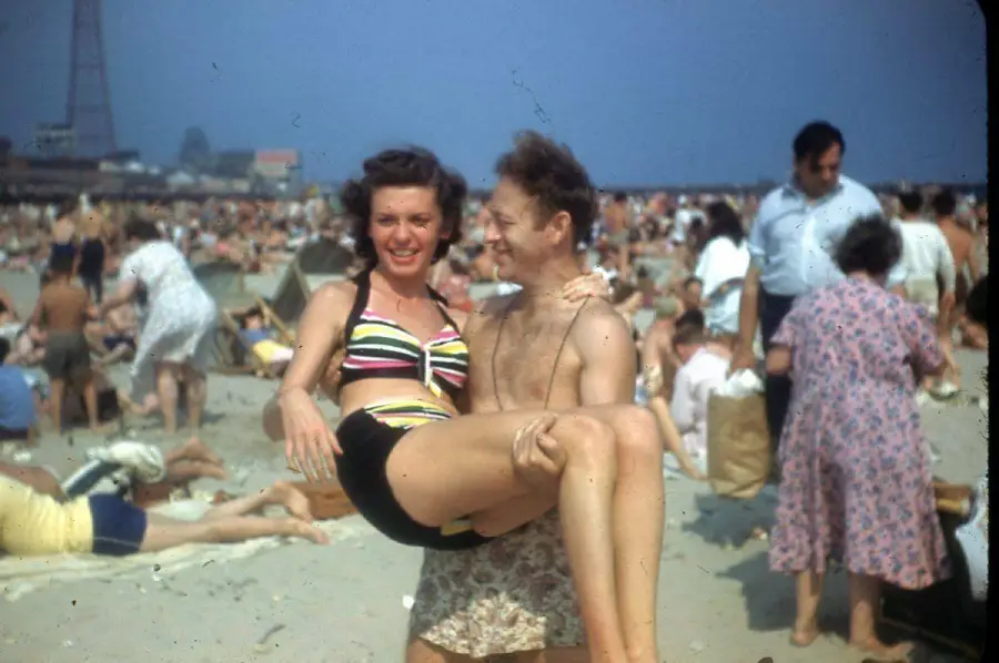 A man in swim trunks lifts a smiling woman in a striped bikini at a crowded beach, with people sunbathing and walking in the background under a clear sky.