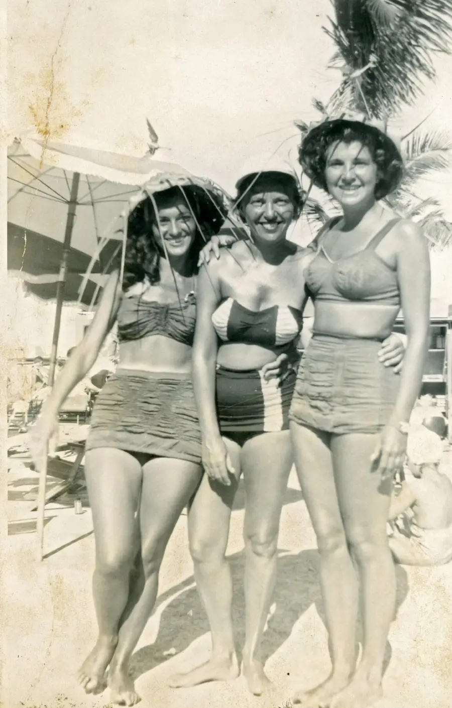 Three women in vintage swimsuits smile and pose together on a sunny beach, with palm trees, umbrellas, and people relaxing in the background. The photo is in black and white.