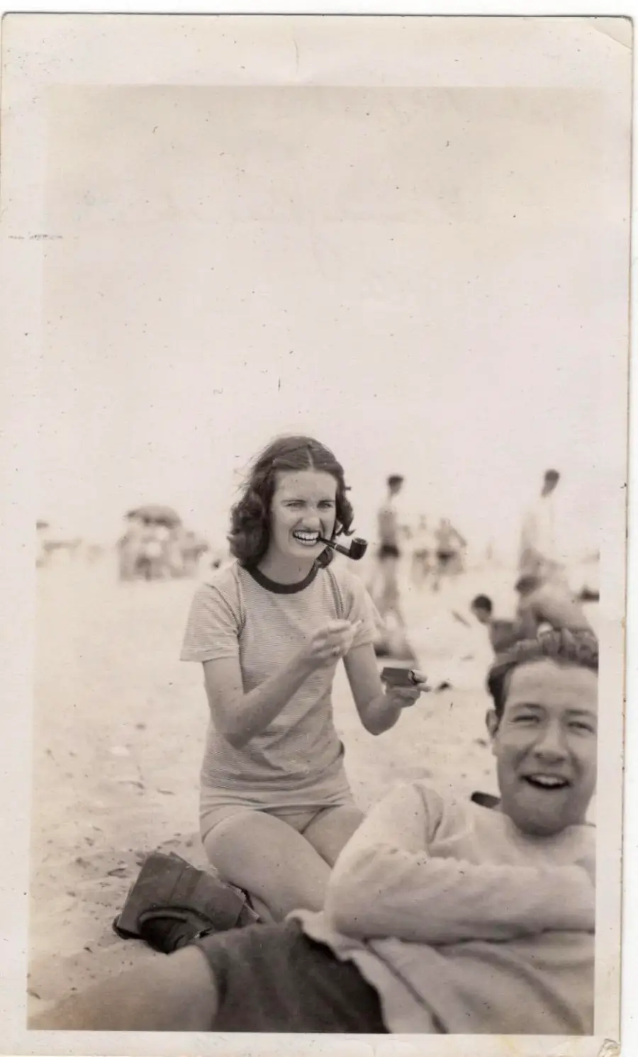 A woman sits on the beach, smiling and holding a pipe in her mouth, while a man lies in front of her, also smiling. People are visible in the blurred background. The photo is black and white.