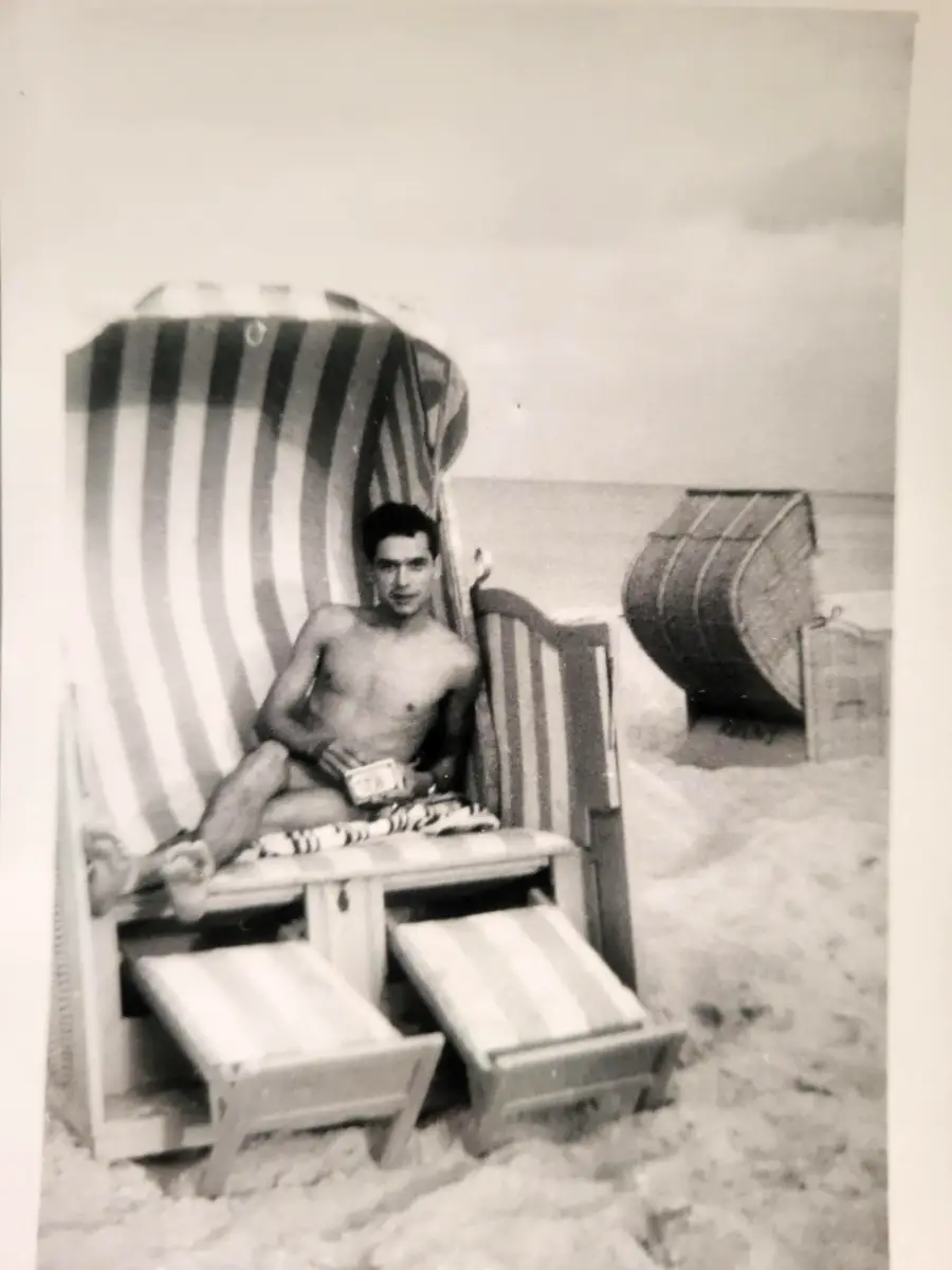 A man sits in a striped beach chair on a sandy beach, with another empty beach chair in the background. The photo is black and white, giving it a vintage feel.
