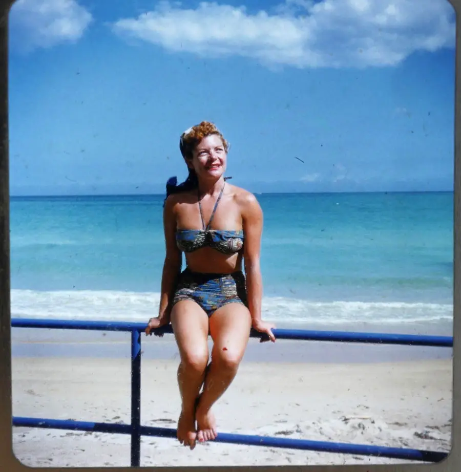 Woman in a vintage bikini sits on a blue railing at the beach, smiling with the ocean and a partly cloudy sky in the background.