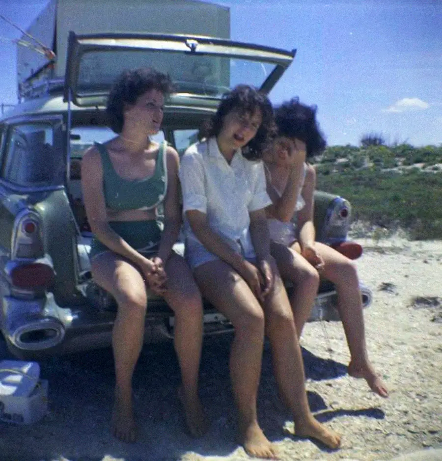 Three women sit barefoot on the open tailgate of a vintage car at a beach, with a cooler nearby and grassy dunes in the background under a blue sky.
