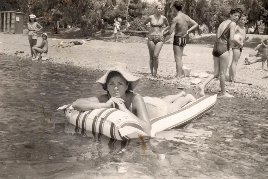 A woman in a wide-brimmed hat lies on an inflatable raft in shallow water near a beach, looking at the camera. Several people stand and relax on the rocky shore in the background.