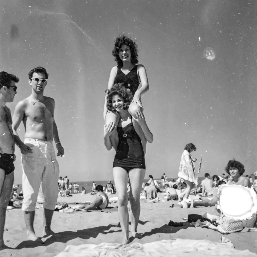 Two women in swimsuits pose, one sitting on the other’s shoulders, on a crowded beach. Two men stand nearby. People are relaxing and playing in the sand under a clear sky. The photo appears to be from the mid-20th century.