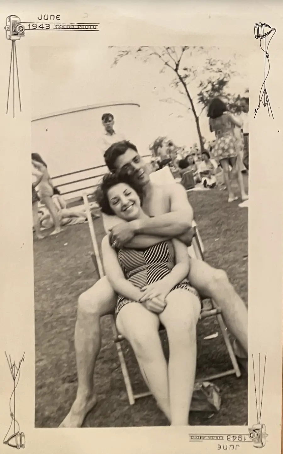 A young couple in vintage swimsuits sits on a lawn chair at a park or beach in June 1943. The man hugs the smiling woman from behind. Other people and trees are visible in the background. Hand-drawn cameras border the photo.