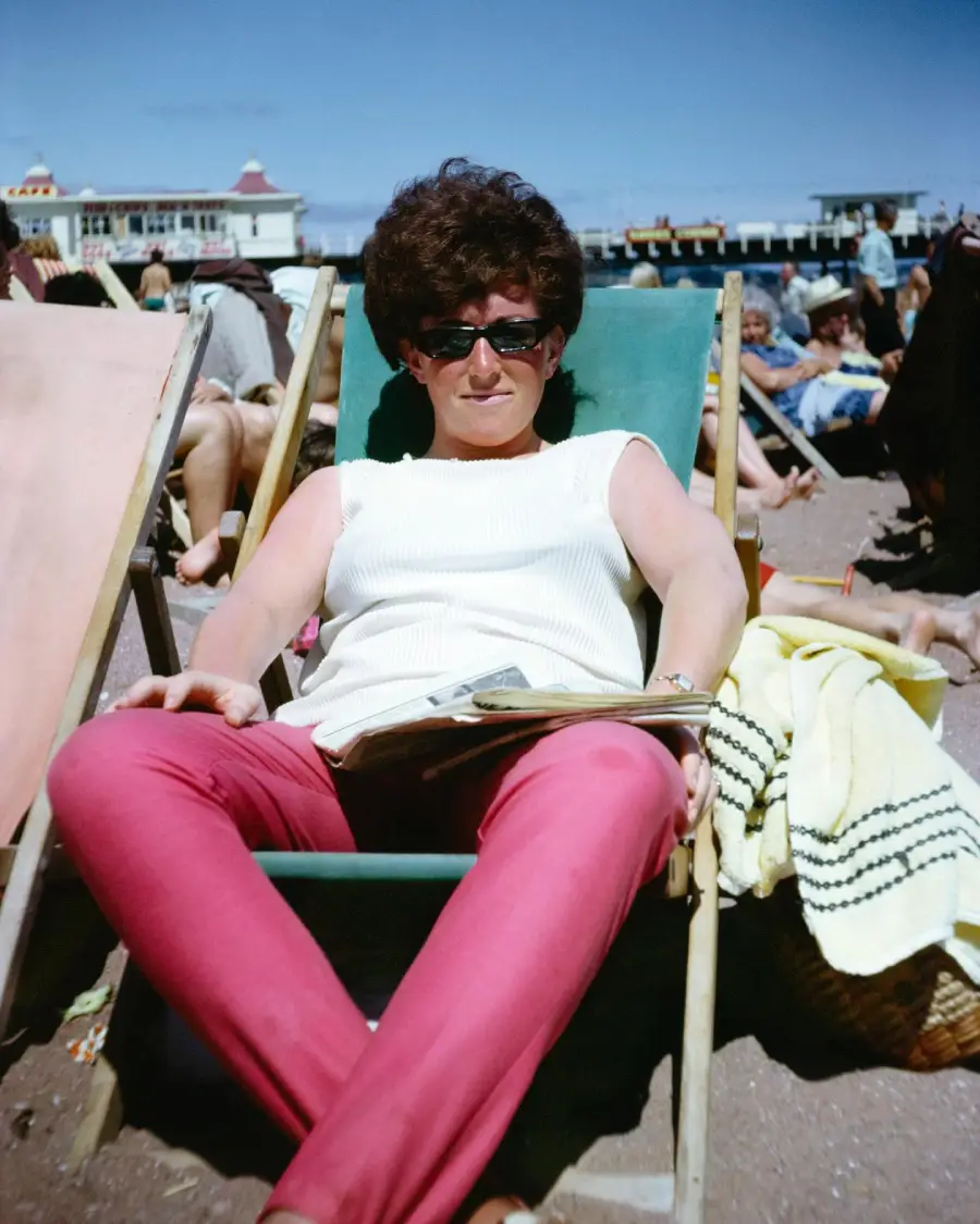 A woman wearing sunglasses, a white sleeveless top, and red pants relaxes on a deck chair at a beach, holding a magazine. People and a pier are visible in the sunny background.