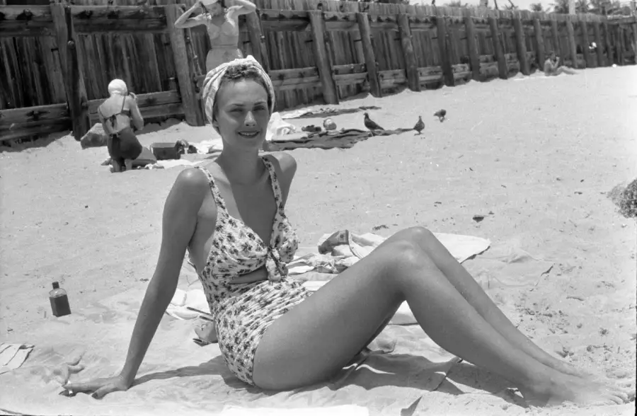 A woman in a vintage swimsuit and headscarf sits on a towel at a sandy beach. A wooden fence, other beachgoers, and birds are visible in the background.