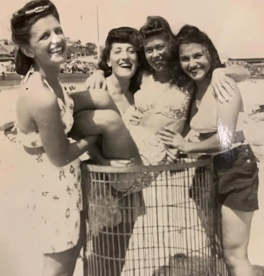 Four women in vintage swimsuits smile and pose together on a beach, with one woman playfully sitting in a large wire trash can as the others gather around her, enjoying a fun moment in the sun.