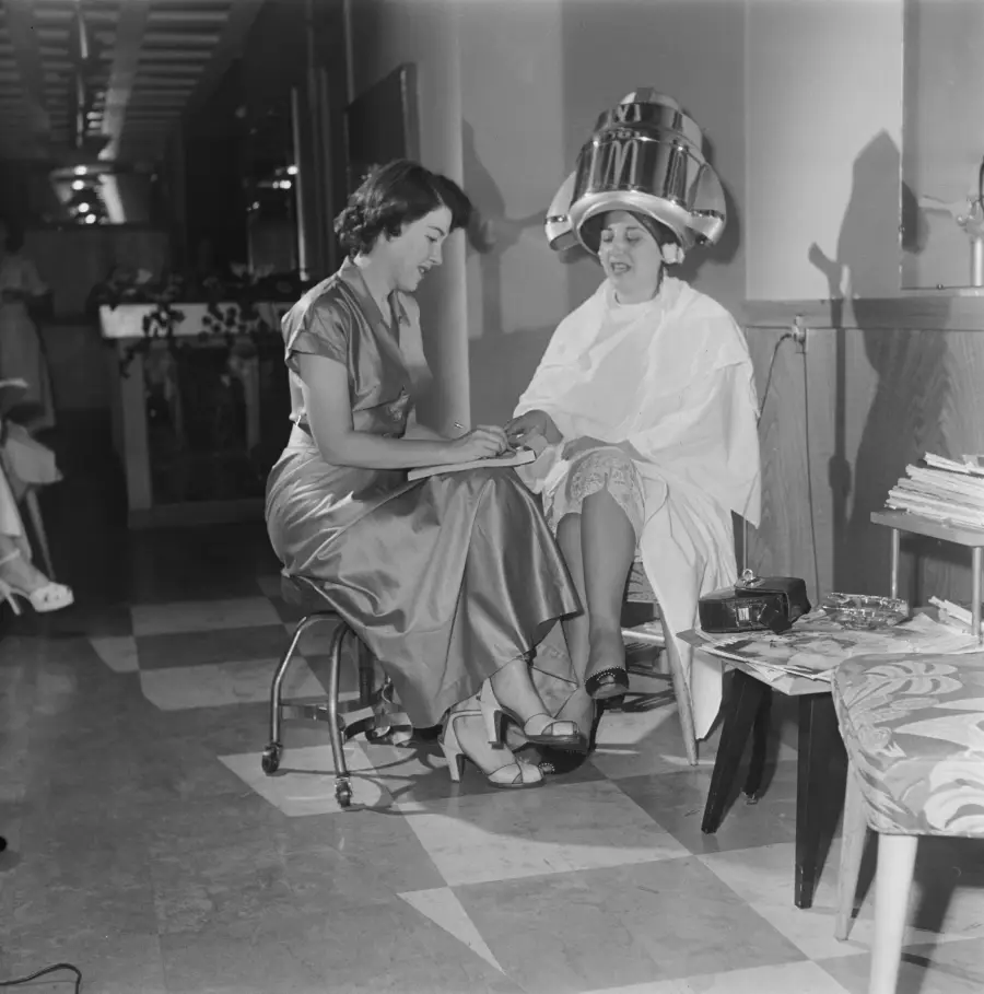 A woman gets a manicure while seated under a hair dryer at a vintage salon. She wears a cape, and the manicurist is in a shiny dress. Magazines and a phone are on a nearby table.