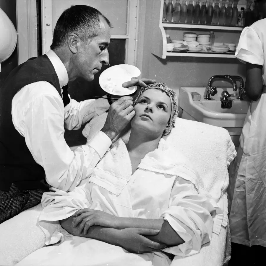 A man applies makeup to a reclining woman in a beauty salon. She wears a robe and head covering, while he holds a palette and focuses on her face. Another person stands at a nearby sink in the background.