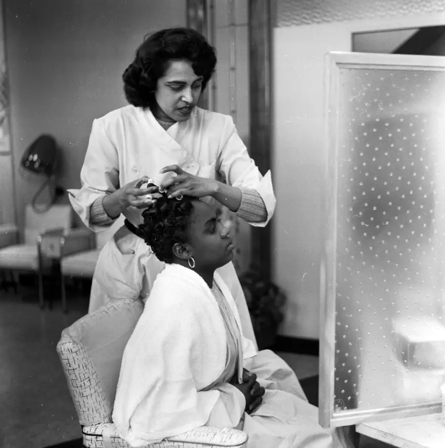 A hairstylist stands behind a seated woman, styling her hair in a salon. The woman wears a robe and faces a mirror, while the background shows salon chairs and a vintage hair dryer.