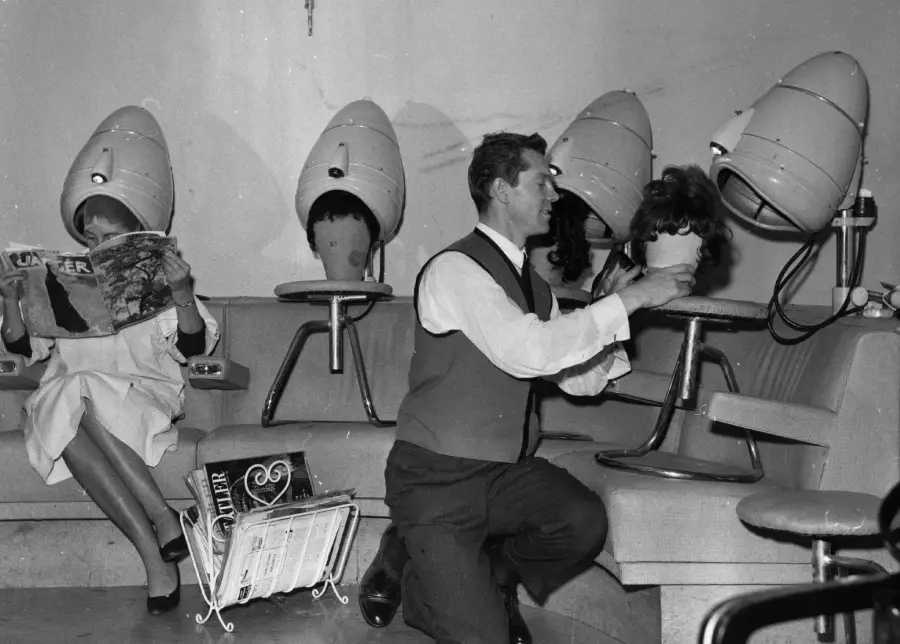 A man styles a woman’s hair under a hood dryer in a vintage salon, while another woman sits beside them reading a magazine, both with hair under dryers. Magazines are scattered on the floor.